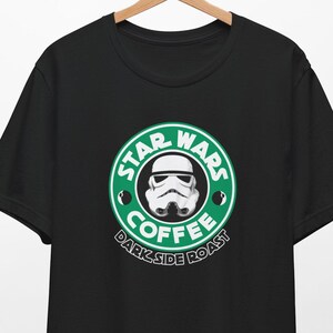 Op de afbeelding: Zwart T-shirt met een Star Wars-geïnspireerd ontwerp. Het ontwerp is een parodie op een koffiehuislogo, met een Stormtrooper-helm in een groene cirkel. De tekst luidt "STAR WARS COFFEE DARK SIDE ROAST".