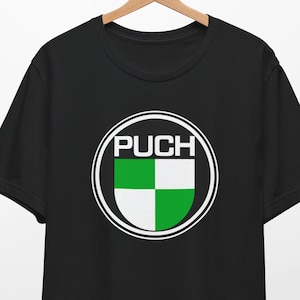 Op de afbeelding: Zwart T-shirt met een wit cirkelvormig logo met het woord "PUCH" in witte letters en een groen-wit geruit patroon in de cirkel.