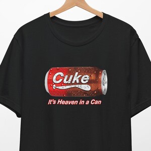 May include: Black t-shirt with a graphic of a red and white can of soda with the word "CUKe" printed on it. The text "It's Heaven in a Can" is printed below the can.