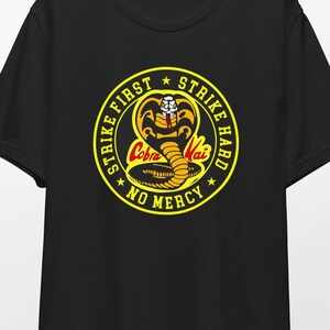 May include: Black t-shirt featuring a yellow and red Cobra Kai logo with the words "Strike First, Strike Hard, No Mercy." The logo depicts a cobra with the words "Cobra Kai" in the center. The shirt is hanging on a wooden hanger.