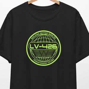 May include: Black t-shirt with a neon green graphic. The graphic is a circular design with the text "BUILDING BETTER WORLDS" and "LV-426". Below the text is the Weyland Corp logo and the words "TERRAFORMING COLONY". The shirt is hanging on a wooden hanger.