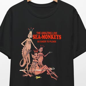 Könnte beinhalten: Schwarzes T-Shirt mit einem Grafikdruck von drei Cartoon-Seemonster, die Kronen tragen. Der Text "THE AMAZING LIVE SEA-MONKEYS SO EAGER TO PLEASE" ist in Rot auf dem Shirt gedruckt.