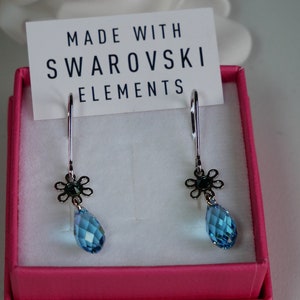 May include: A pair of blue teardrop earrings with a silver flower charm. The earrings are in a pink box with a white card that reads "Made with Swarovski Elements".