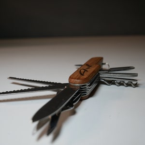 Könnte beinhalten: Ein Multifunktions-Taschenmesser mit einem Holzgriff, der mit den Initialen "A & L" graviert ist. Das Messer verfügt über eine Vielzahl von Werkzeugen, darunter eine Säge, einen Korkenzieher und einen Schraubendreher.