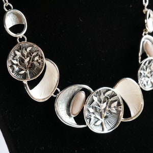 May include: A silver necklace with a tree of life design. The necklace features several round and crescent-shaped pendants with a textured white finish.