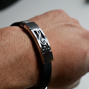 May include: A black leather bracelet with a silver metal plate featuring a treble clef design.