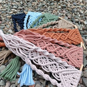 May include: A set of nine macrame wall hangings in various colours, including blue, green, brown, and pink. Each hanging features a unique pattern and has a tassel at the bottom.