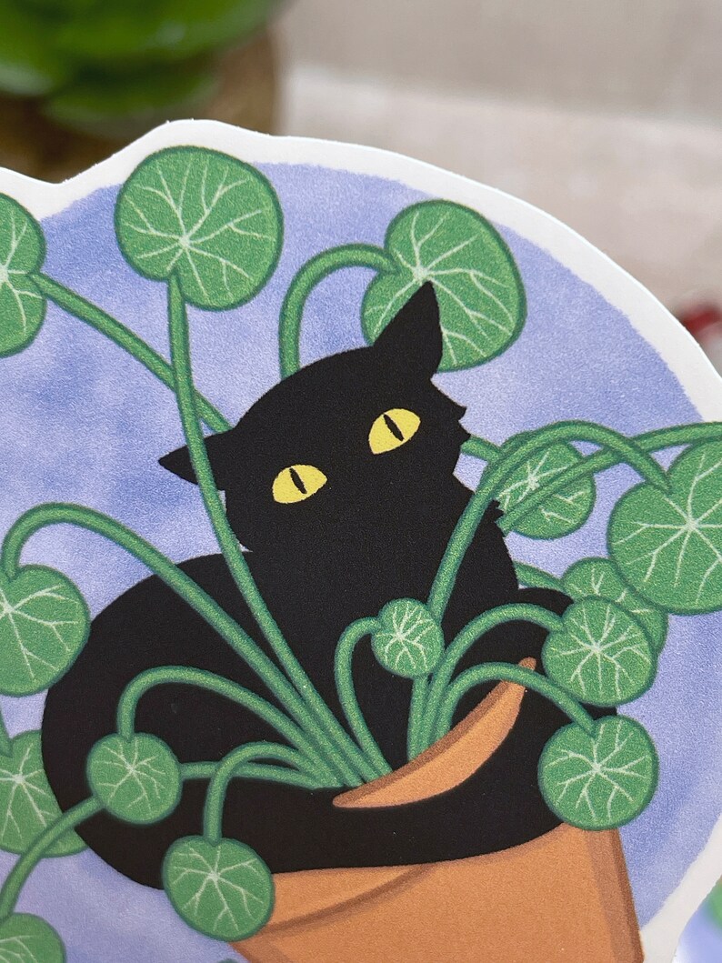 May include: A black cat with yellow eyes is nestled in a pot of green leafy plants. The cat is partially hidden by the leaves, with only its head and part of its body visible. The pot is brown and has a light blue background.