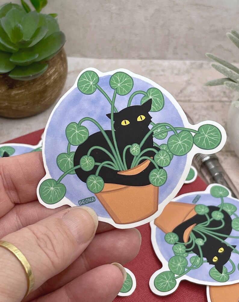 May include: A black cat is hiding in a pot of green leafy plants. The cat has yellow eyes and is sitting in the pot. The pot is brown and has a white rim. The background is a light blue circle with a white outline.