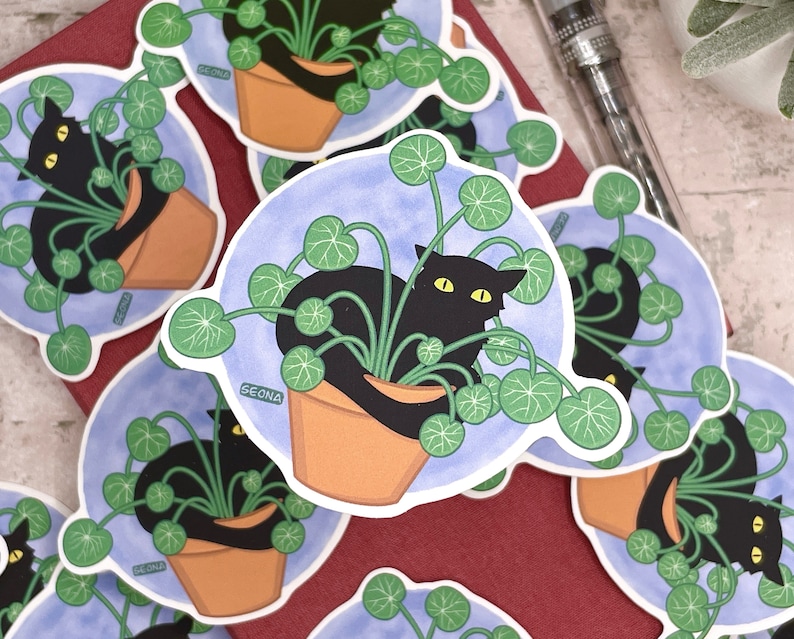 May include: A set of stickers featuring a black cat curled up in a pot of green plants. The stickers have a blue background with white text that says "Seona".