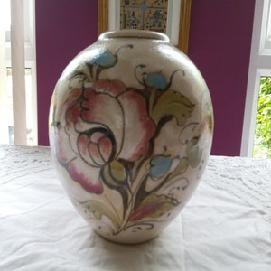 May include: A cream-coloured, bulbous ceramic vase with a hand-painted floral design. The flowers are pink and red, with green leaves and blue accents. The vase is on a white tablecloth.