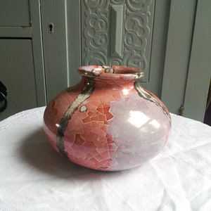 May include: A round ceramic vase with a crackle glaze finish. The vase features a color palette of pink, lavender, and gray. The vase is sitting on a white surface.
