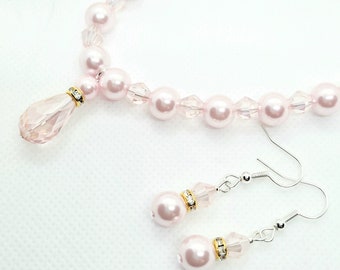 Puede incluir: Un collar de perlas rosas con un colgante de cristal y pendientes a juego de perlas rosas con detalles de cristal.