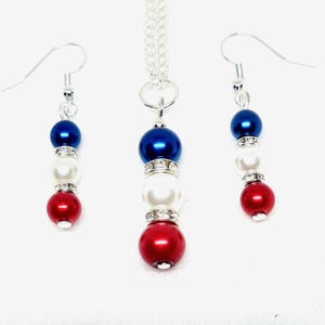 May include: A jewelry set featuring a necklace and earrings. The necklace has a silver chain with a pendant of three beads: blue, white, and red. The earrings mirror the pendant design. Each bead is separated by a rhinestone spacer.