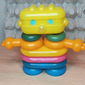 May include: A colourful plastic toy robot with a yellow head, green and orange rings, and blue feet.