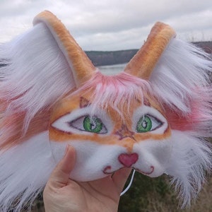 May include: A white and orange furry cat mask with green eyes and a pink nose. The mask has a white fur trim around the edges.