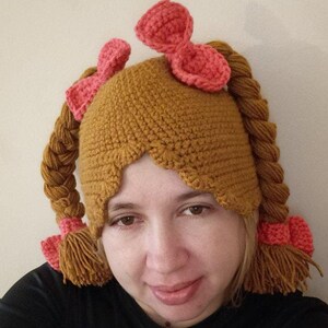 May include: A brown crocheted hat with two long braids and pink bows. The hat is worn by a person with brown hair and eyes.