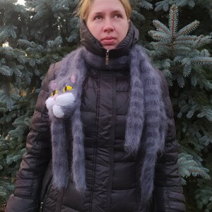 May include: A woman wearing a black puffer jacket with a gray fuzzy scarf featuring a cat face appliqu&eacute;. The scarf is worn as a neck warmer and has a long, flowing design.
