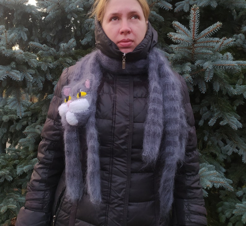 May include: A woman wearing a black puffer jacket with a gray fuzzy scarf featuring a cat face appliqu&eacute;. The scarf is worn as a neck warmer and has a long, flowing design.