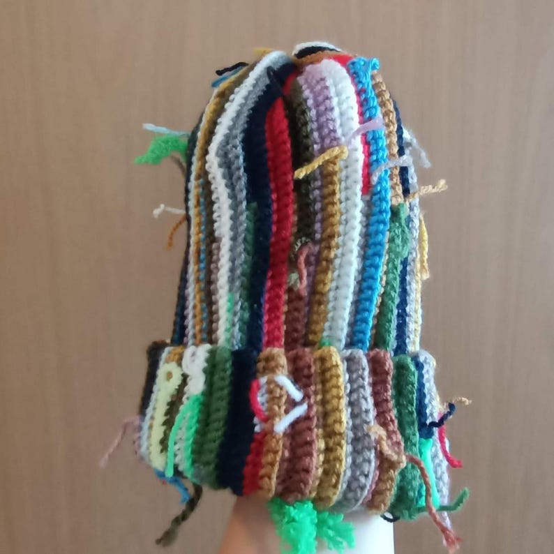 Op de afbeelding: Een handgemaakte gehaakte beanie in diverse kleuren, waaronder groen, blauw, rood en bruin. De muts heeft een omgeslagen rand en heeft meerdere strengen garen die uit de bovenkant en de rand steken. Een uniek, kleurrijk accessoire.