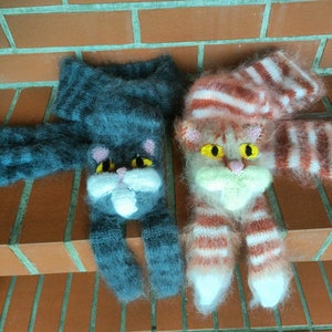 May include: Two knitted cat-shaped fingerless gloves. One is gray with pink ears and nose, the other is pink and white with orange stripes and yellow eyes.