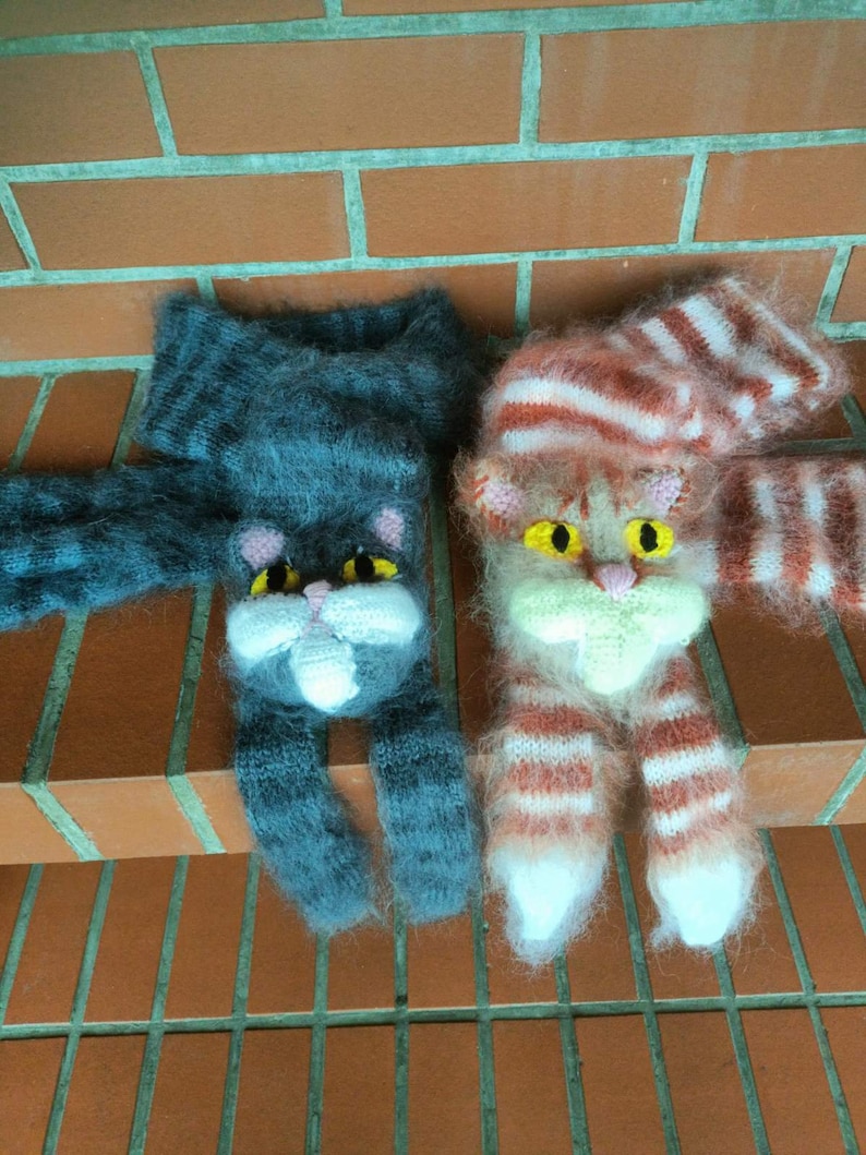May include: Two knitted cat-shaped fingerless gloves. One is gray with pink ears and nose, the other is pink and white with orange stripes and yellow eyes.