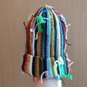 Op de afbeelding: Een handgemaakte beanie-muts met een kleurrijk gestreept ontwerp. De muts heeft verticale strepen in verschillende kleuren, waaronder rood, blauw, groen en geel. De muts heeft een omgeslagen rand en verschillende garenuiteinden die van de bovenkant en de rand hangen.