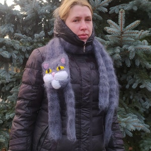 May include: A woman wearing a black jacket with a gray scarf featuring a knitted cat head. The cat has yellow eyes and a white face.