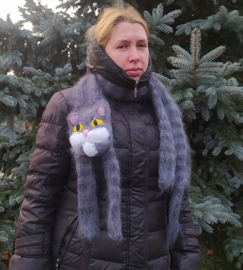 May include: A woman wearing a black jacket with a gray scarf featuring a knitted cat head. The cat has yellow eyes and a white face.