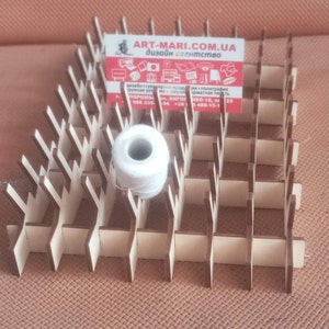 May include: A wooden thread organiser with a white spool of thread and a business card. The organiser is made of interlocking wooden pieces, creating a grid-like structure. The business card has text and a logo.