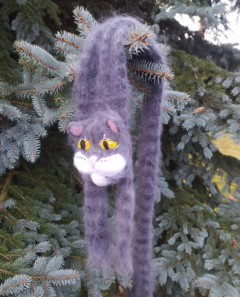 May include: A gray and white knitted scarf with a cat face design. The cat has yellow eyes and a white nose and whiskers.