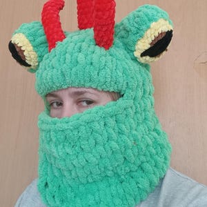 May include: Green knitted frog hat with red antennas and black eyes. The hat has a face opening and a long neck covering.