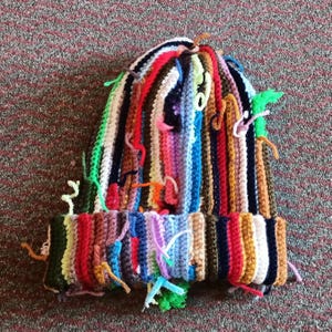 Op de afbeelding: Een kleurrijke, handgemaakte gehaakte beanie. De hoed heeft verticale strepen in verschillende kleuren, waaronder rood, blauw, groen en geel. De hoed heeft een omgeslagen rand en verschillende losse garenuiteinden. Een uniek accessoire.