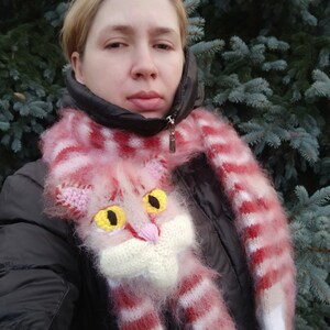 May include: A pink and white striped scarf with a fluffy texture features a knitted cat face with yellow eyes. The scarf is worn by a person standing in front of a green evergreen tree.