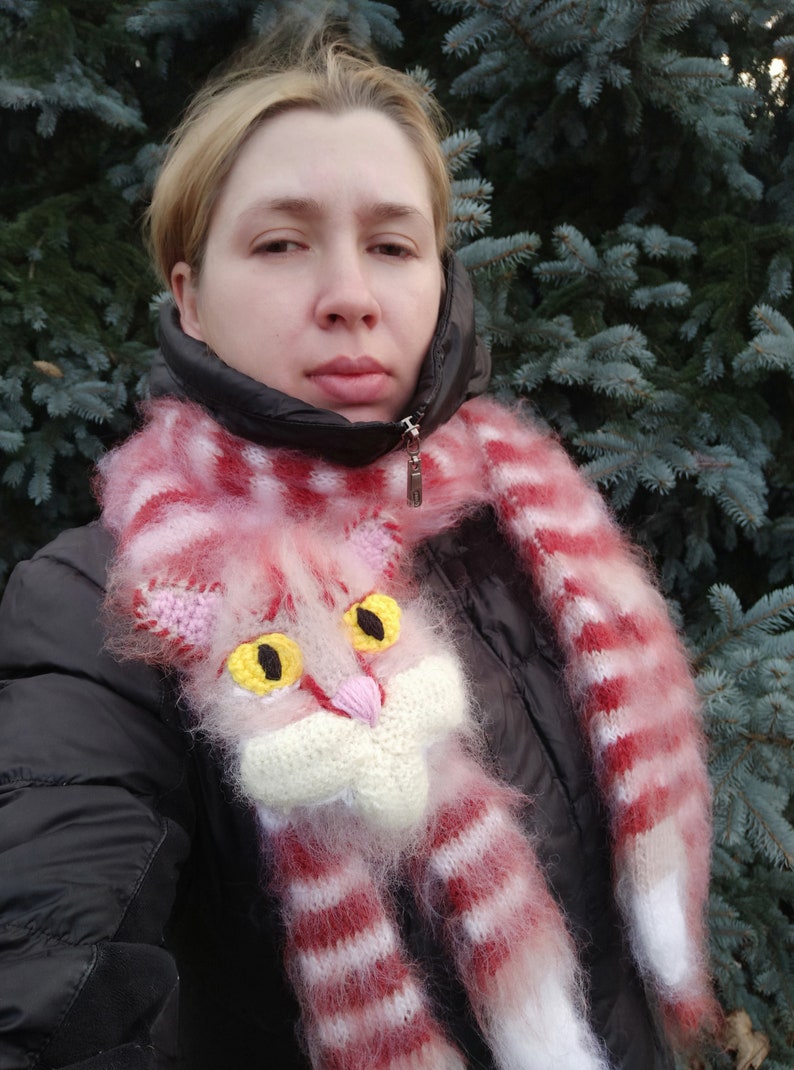 May include: A pink and white striped scarf with a fluffy texture features a knitted cat face with yellow eyes. The scarf is worn by a person standing in front of a green evergreen tree.
