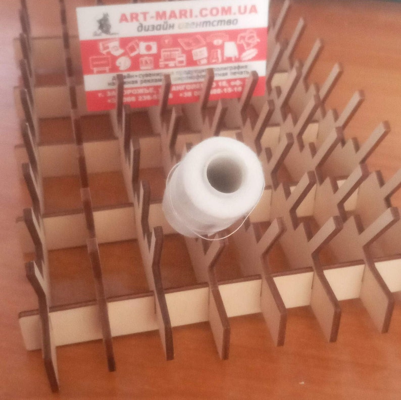 May include: A light brown wooden craft organiser with a grid pattern, holding a white thread spool. The background features a business card with the text "ART-MARI.COM.UA". The organiser is made of thin wooden slats.