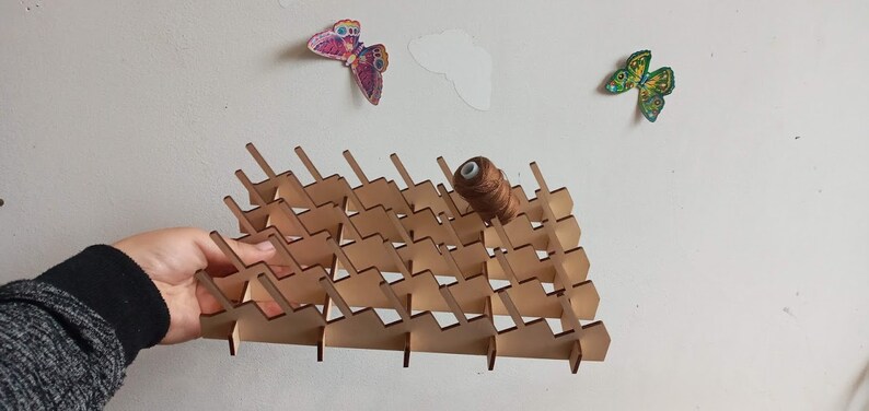 May include: A wooden thread rack with a brown spool of thread on top. The rack has a triangular shape and is made of light brown wood. It has multiple slots for holding spools of thread.