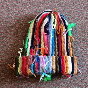 Op de afbeelding: Een kleurrijke, handgemaakte beanie-muts met verticale strepen in verschillende kleuren, waaronder rood, blauw, groen en bruin. De muts heeft een omgeslagen rand en garenuiteinden die van de bovenkant hangen, wat het een textuur geeft.