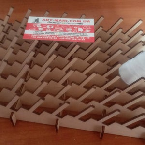May include: A wooden wine glass rack with a distinctive geometric design. The rack is made of light brown wood and features angled slots to hold wine glasses. A white object rests on the rack. A business card is on the rack.