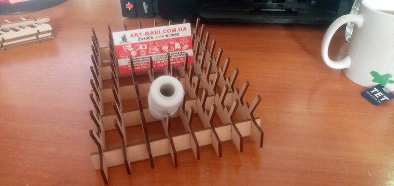 May include: A wooden business card holder with a geometric design, holding a business card with the text "ART-MARI.COM". A roll of white tape sits in the holder. A mug is in the background.