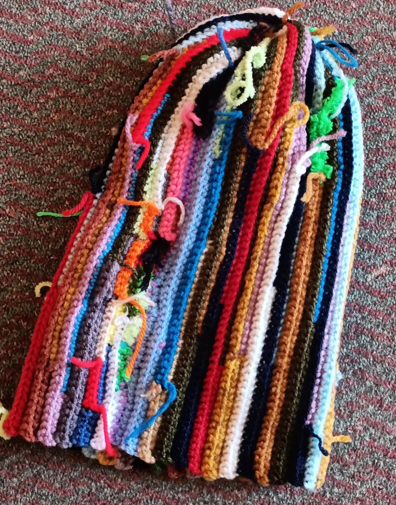 Op de afbeelding: Een handgemaakte, kleurrijke gehaakte muts met verticale strepen in verschillende kleuren, waaronder rood, blauw, groen en bruin. De muts heeft verschillende losse garenuiteinden, wat bijdraagt aan de unieke, handgemaakte uitstraling.