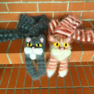 May include: Two knitted cat scarves with fuzzy yarn. One scarf is gray with black stripes and the other is pink with white stripes. Both scarves have embroidered eyes and a white nose.