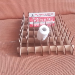 May include: A wooden thread spool organiser with multiple slots, holding a white spool of thread. The organiser has a stepped design and a business card with text. The background is a solid, muted orange.