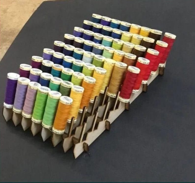 May include: A wooden thread rack with multiple rows of spools of thread in various colors, including red, orange, yellow, green, blue, purple, and black.