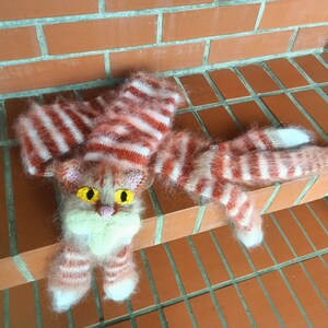 May include: A knitted cat toy with pink and white stripes. The cat has yellow eyes and a pink nose.
