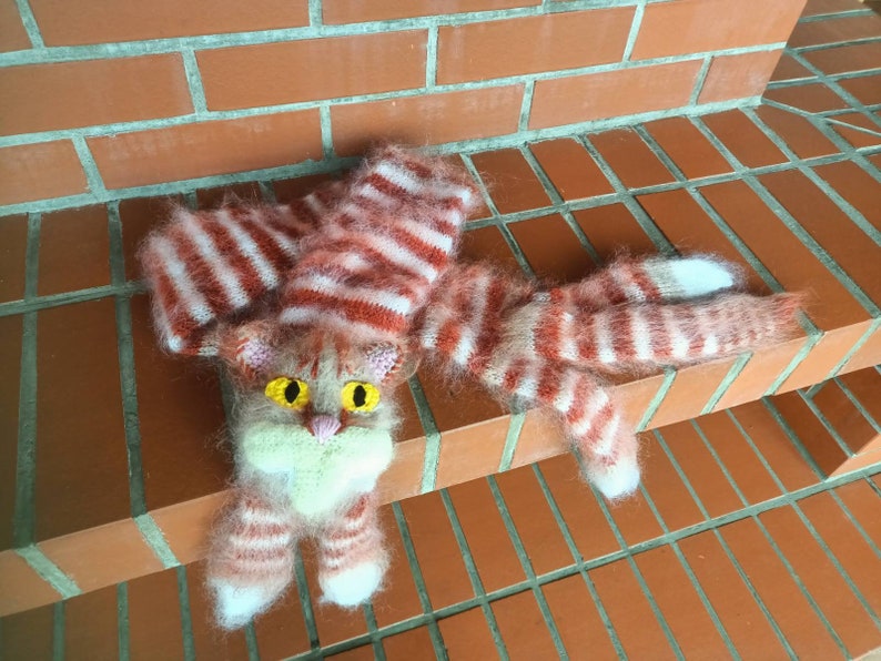 May include: A knitted cat toy with pink and white stripes. The cat has yellow eyes and a pink nose.
