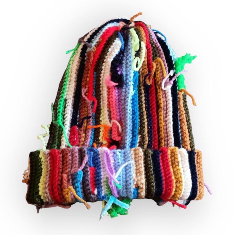 Op de afbeelding: Een kleurrijke, handgemaakte beanie-muts met verticale strepen in verschillende kleuren, waaronder rood, groen, blauw en geel. De muts heeft een omgeslagen rand en losse garenuiteinden, wat een textuurrijke, artistieke uitstraling geeft. Een uniek accessoire.