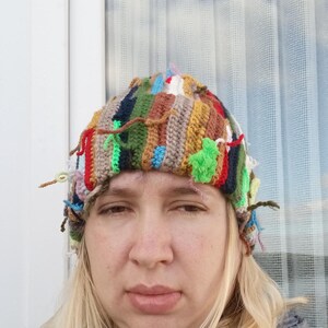 Op de afbeelding: Een kleurrijke, handgemaakte gehaakte hoed met een patchwork-ontwerp. De hoed heeft verschillende kleuren, waaronder rood, groen, blauw en bruin, met getextureerd garen en losse uiteinden. Een uniek, artistiek accessoire.