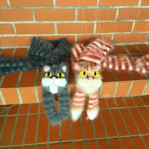 May include: Two knitted cat scarves with fuzzy yarn. One scarf is gray with black stripes and the other is pink with white stripes.