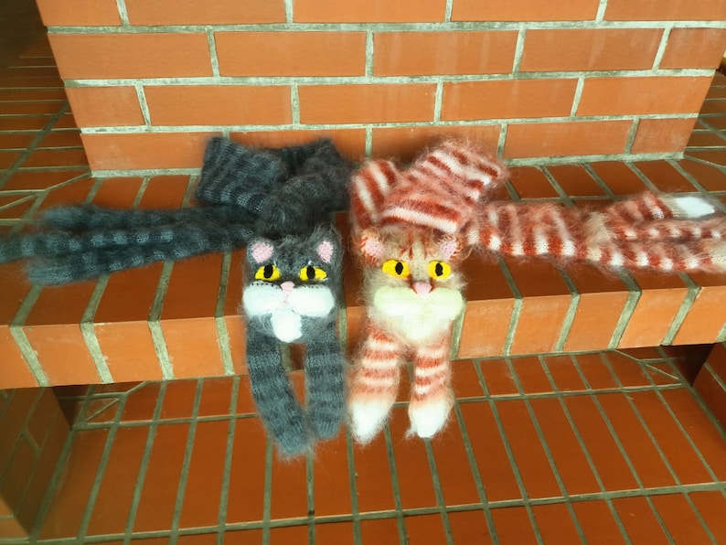 May include: Two knitted cat scarves with fuzzy yarn. One scarf is gray with black stripes and the other is pink with white stripes.